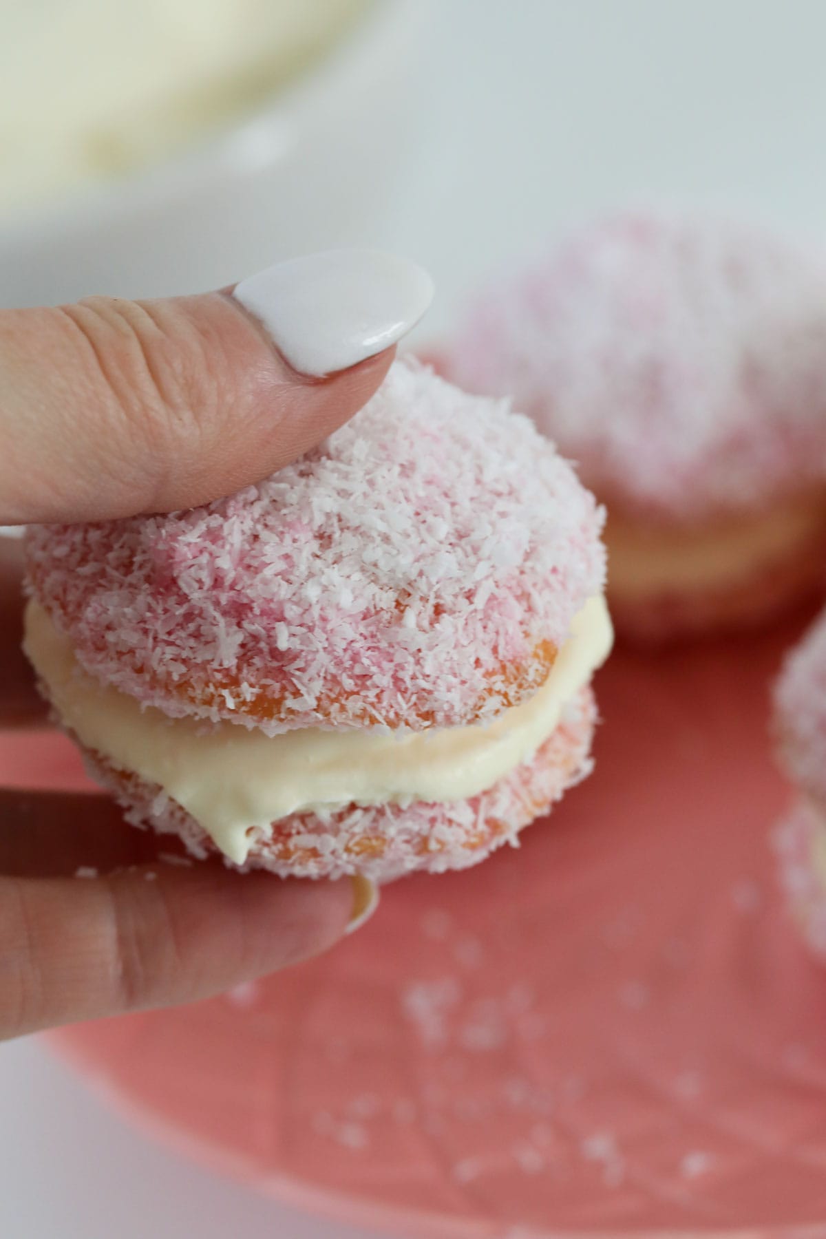 A hand holding a small pink sponge, coated in coconut and sandwiched together with cream.