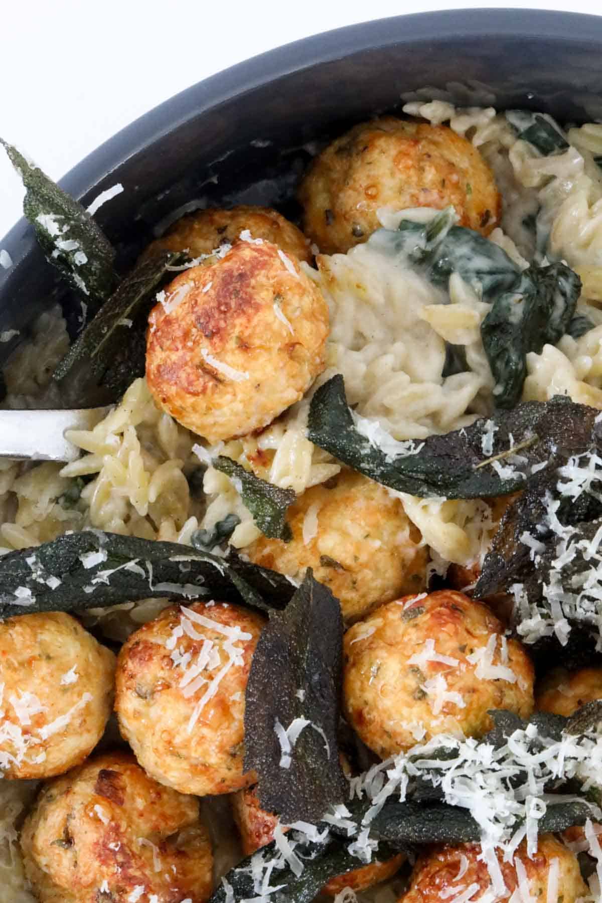 A close up of chicken meatballs and creamy risoni, with crispy sage leaves and finely grated parmesan on top.
