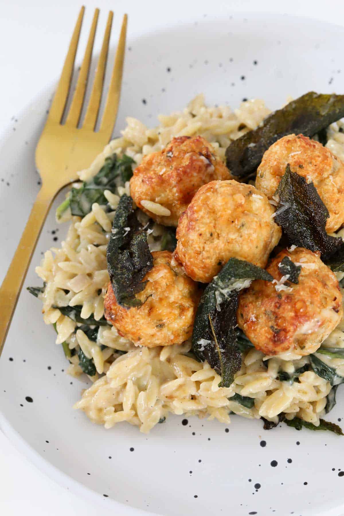 A gold fork placed beside a serve of pasta and chicken balls, sprinkled with crispy sage leaves.