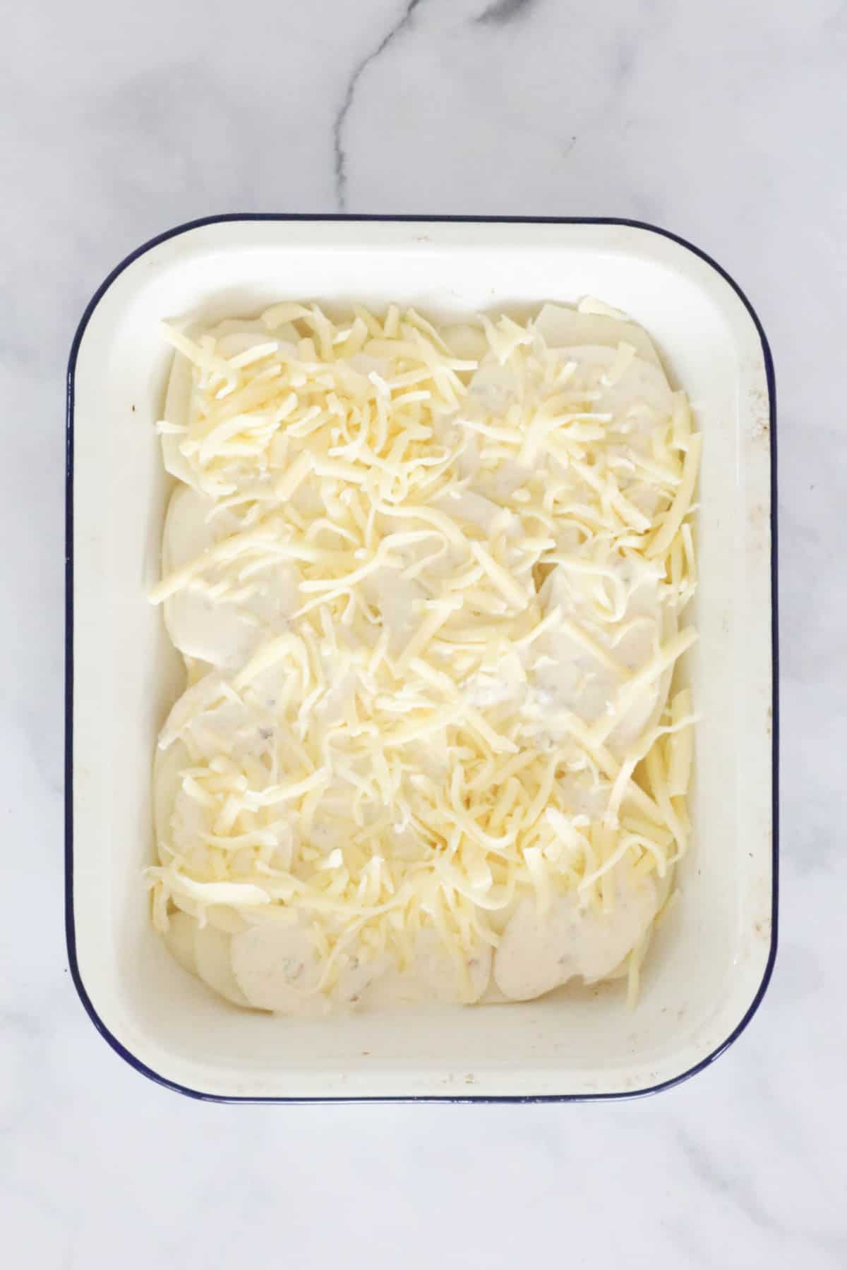 A layer of sliced potato, and cream mixture with shredded cheese sprinkled over top, in a baking dish.
