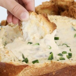 A hand putting a piece of crusty bread into a gooey jalapeno popper cob loaf dip.