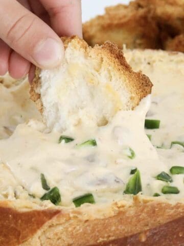 A hand putting a piece of crusty bread into a gooey jalapeno popper cob loaf dip.