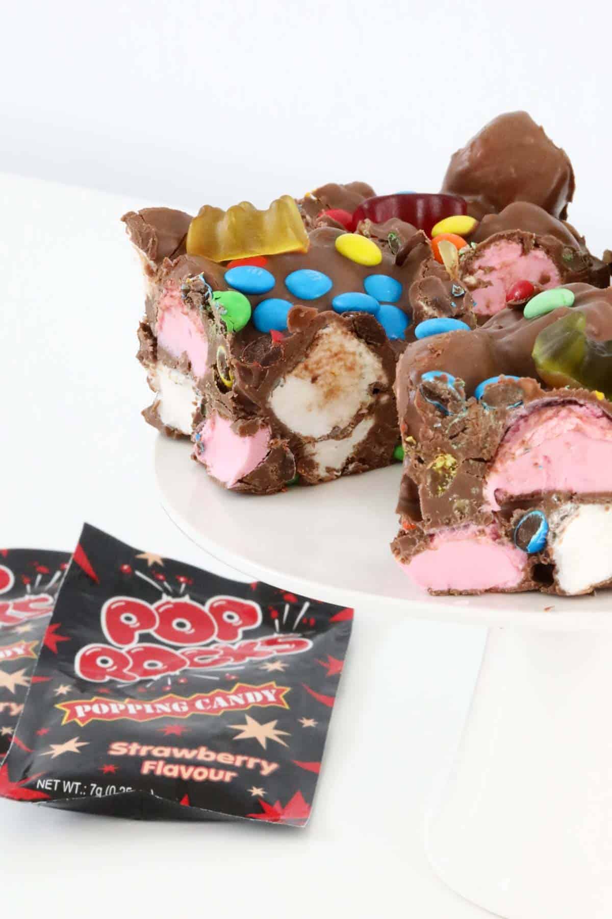 Slices of popping candy rocky road on a white cake stand, with packets of popping candy to the left side.