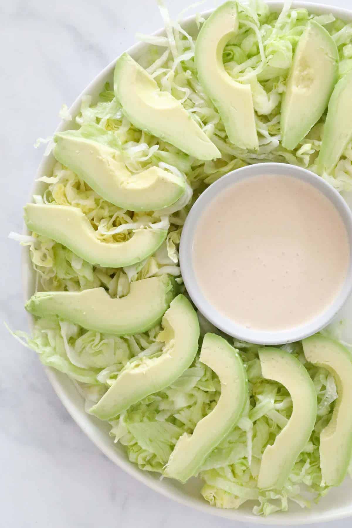 The ramekin of cocktail sauce placed on a serving platter surrounded by shredded lettuce and sliced avocado.