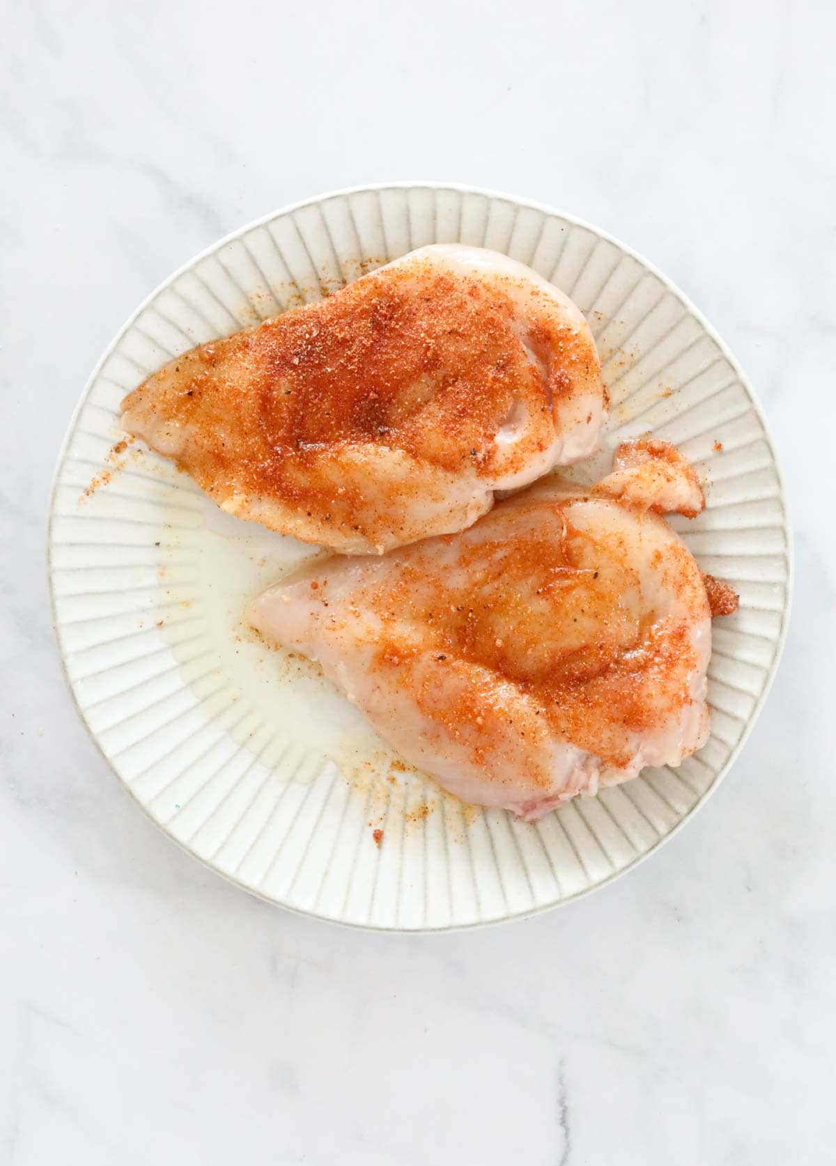 Two raw chicken breasts on a plate, rubbed with the spice mix.