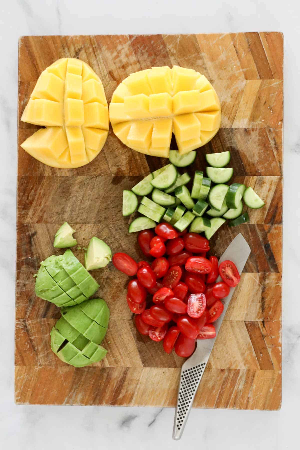 A cubed mango, avocado, cucumber and tomatoes on a wooden chopping board.