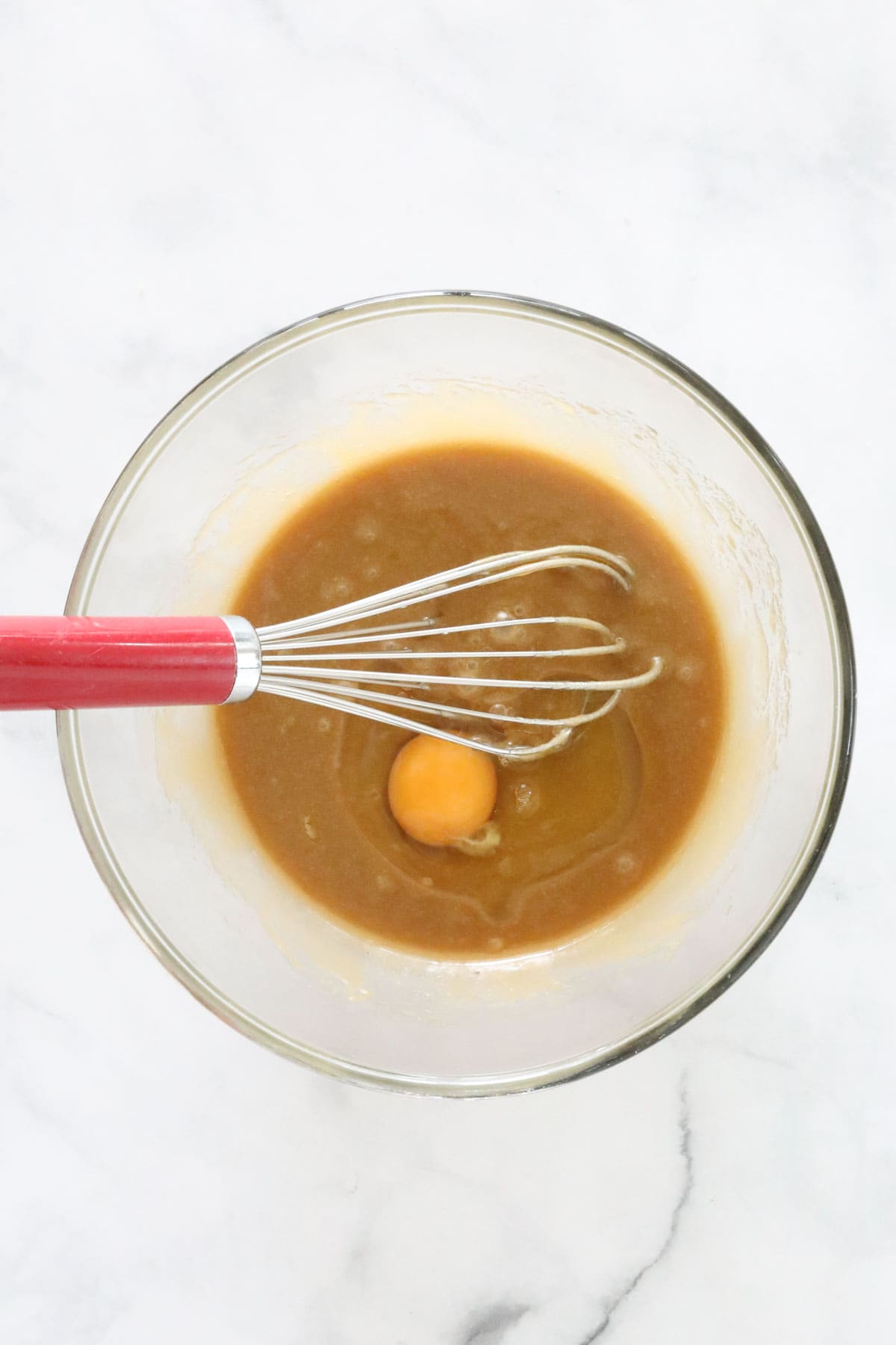 An egg added to the melted butter mixture in a glass bowl with a whisk.