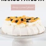 A pavlova decorated with fresh mango slices and blueberries on a cake stand.