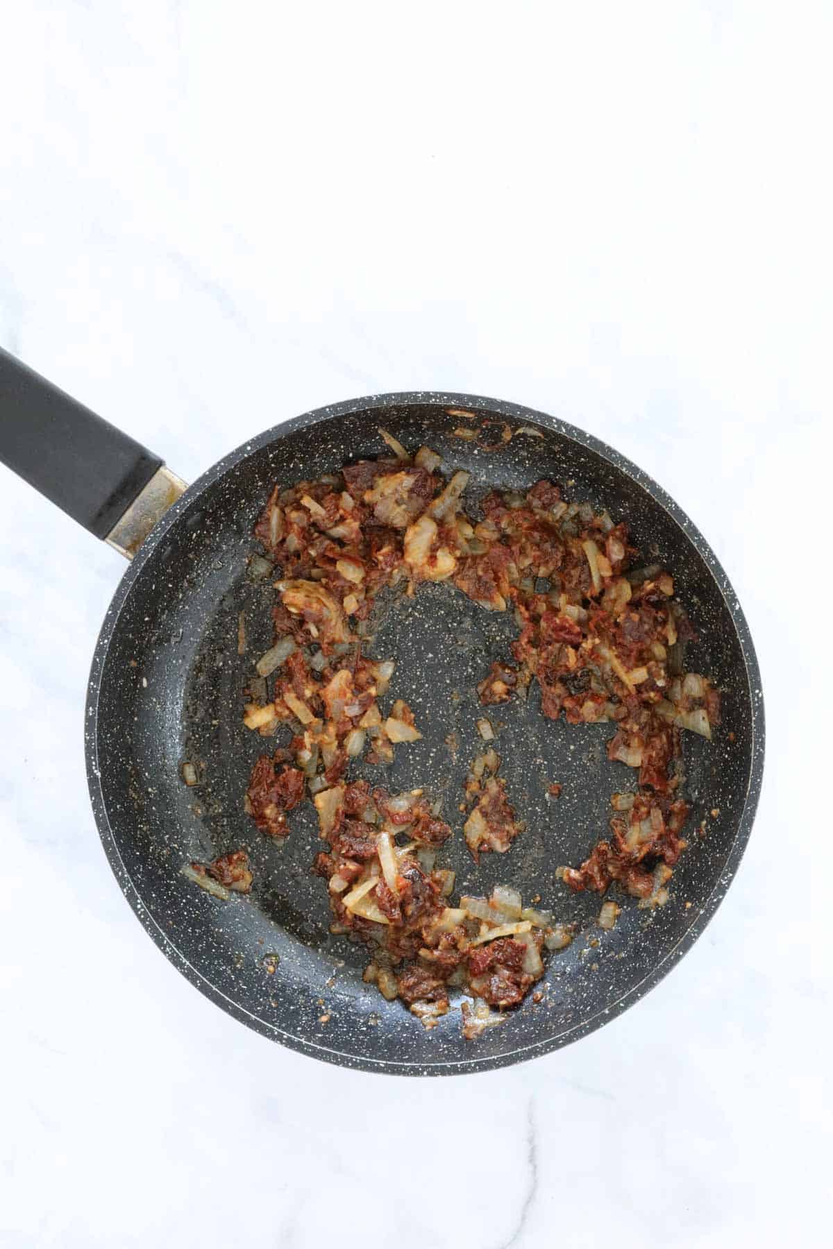 Cooked onion, garlic and sun dried tomatoes in a frying pan.
