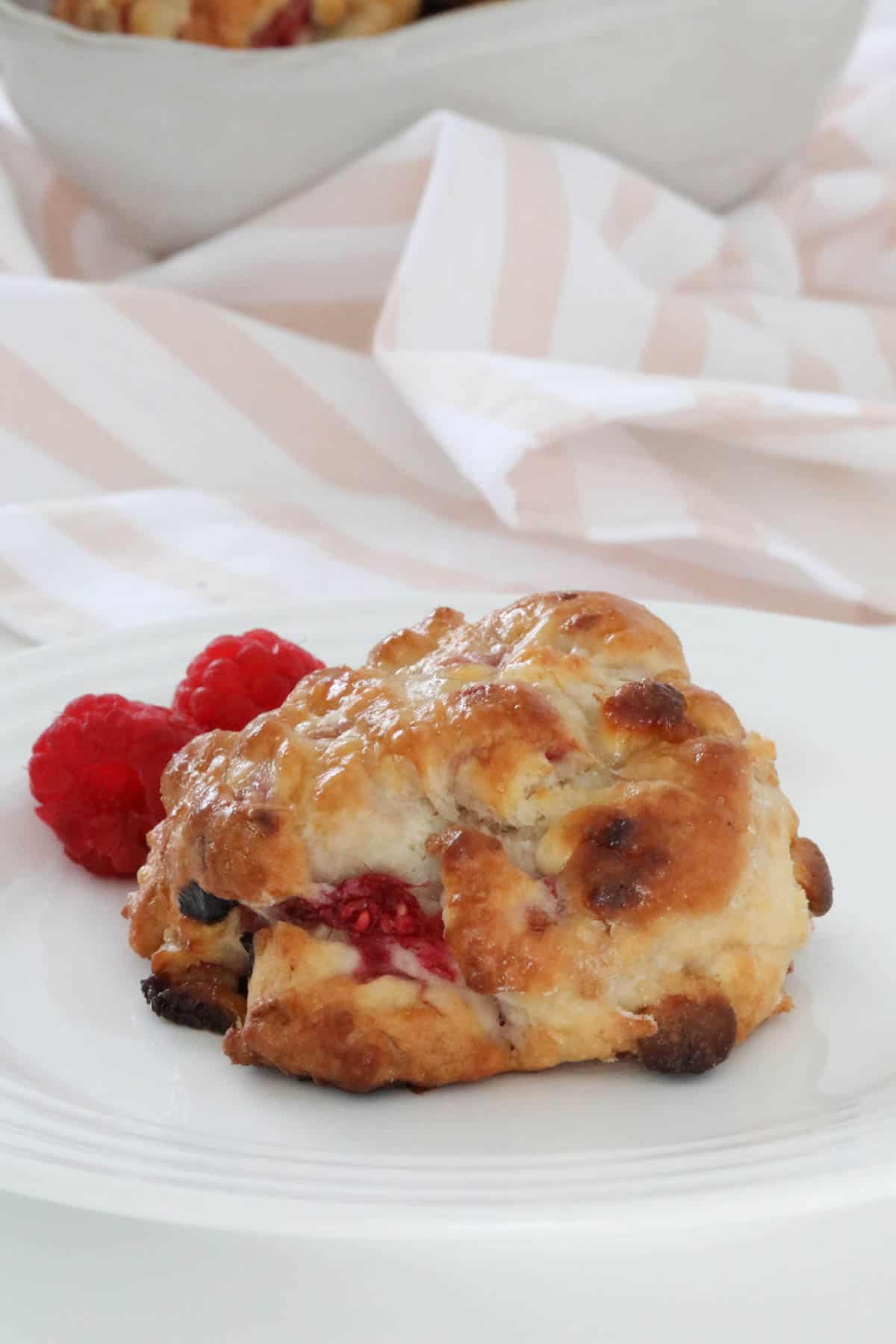 A scone on a plate with two fresh raspberries placed beside it.