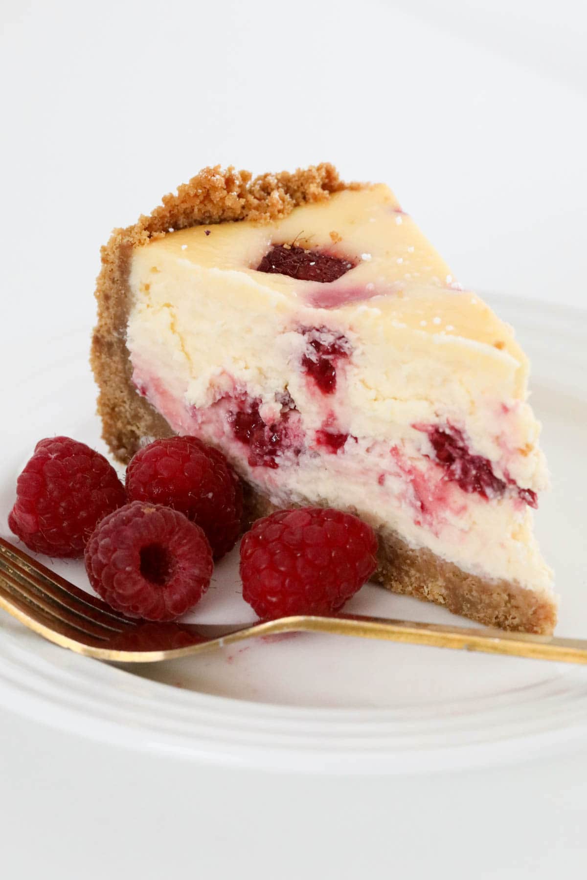 A serve of baked cheesecake with fresh raspberries and a gold fork placed beside it.