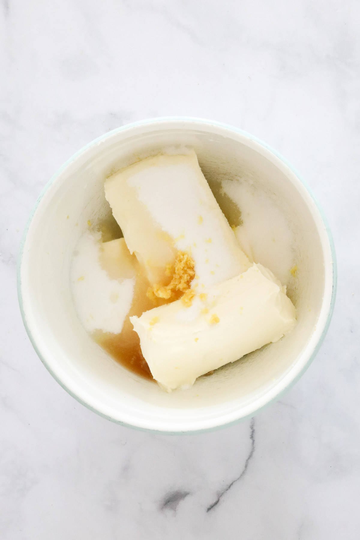 Blocks of cream cheese, sugar, vanilla, lemon juice and zest placed in a large white bowl.