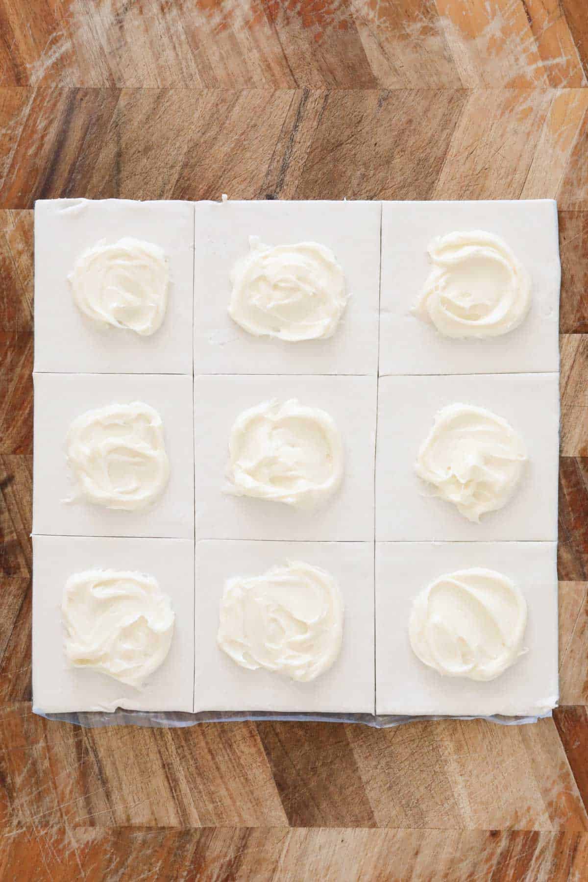 A small spoonful of cream cheese mixture spread on each square of a pastry sheet that has been cut into nine squares.