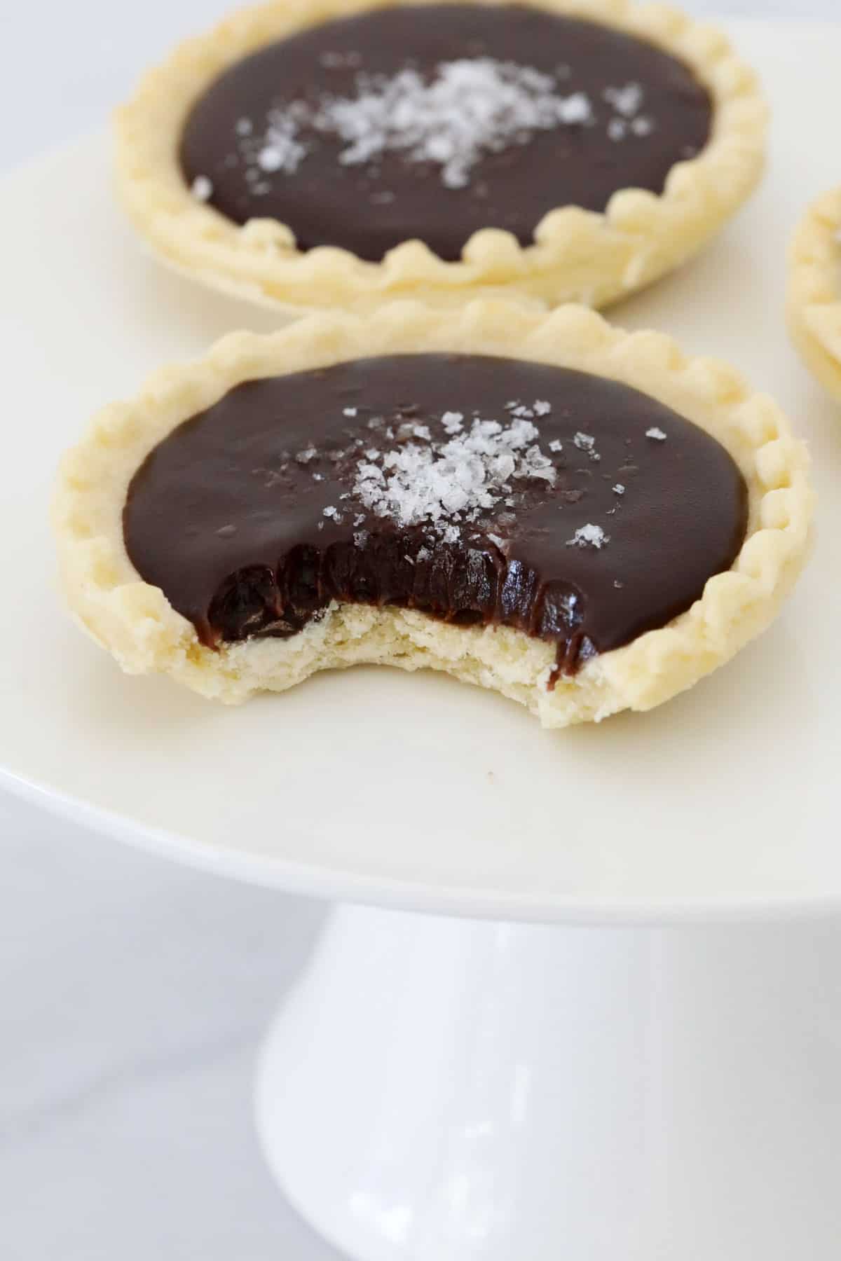 Two chocolate tarts on a white cake stand, one with bite out to show the chocolate ganache texture.