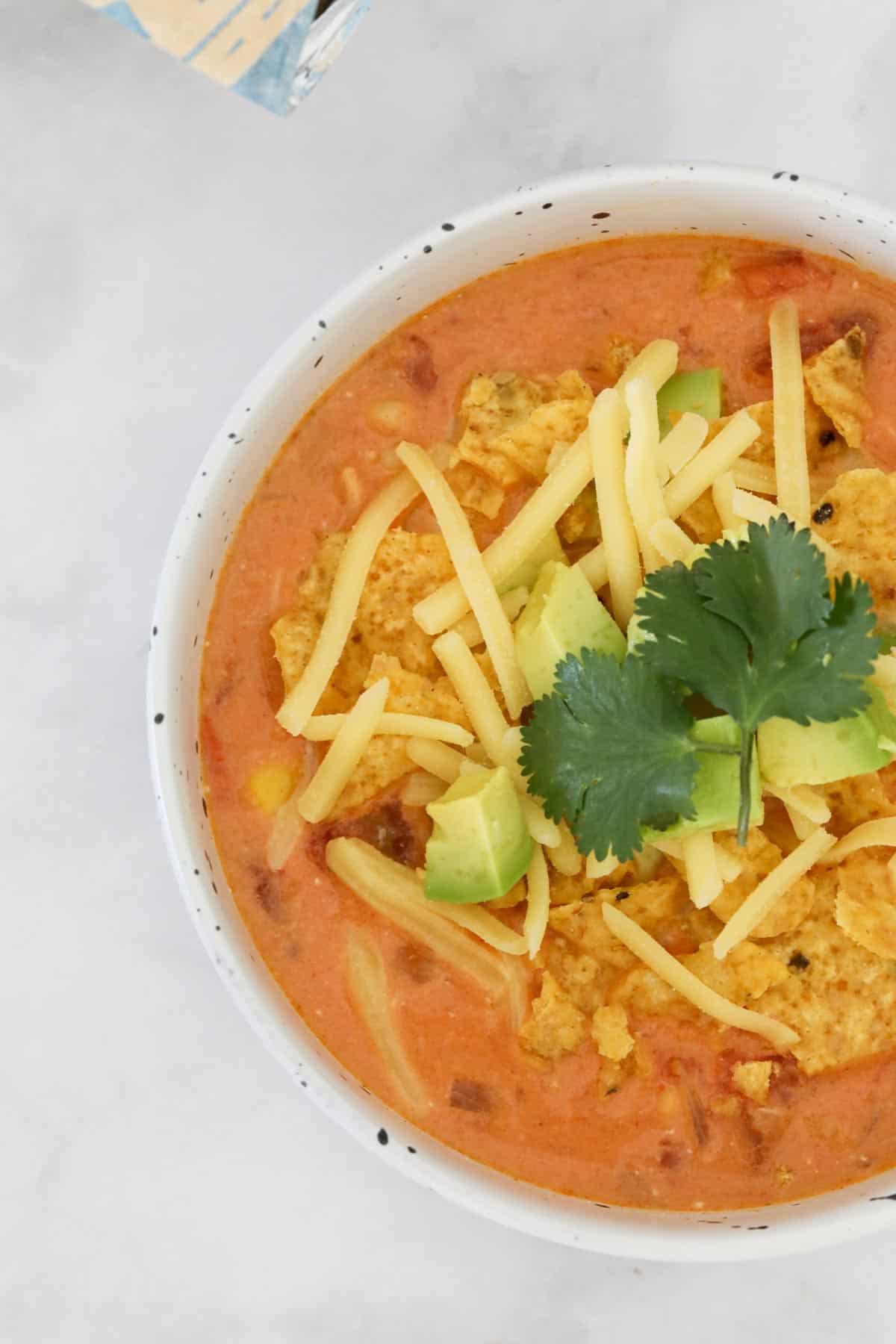 A bowl of chicken taco soup topped with corn chips, grated cheese, avocado chunks and fresh coriander.