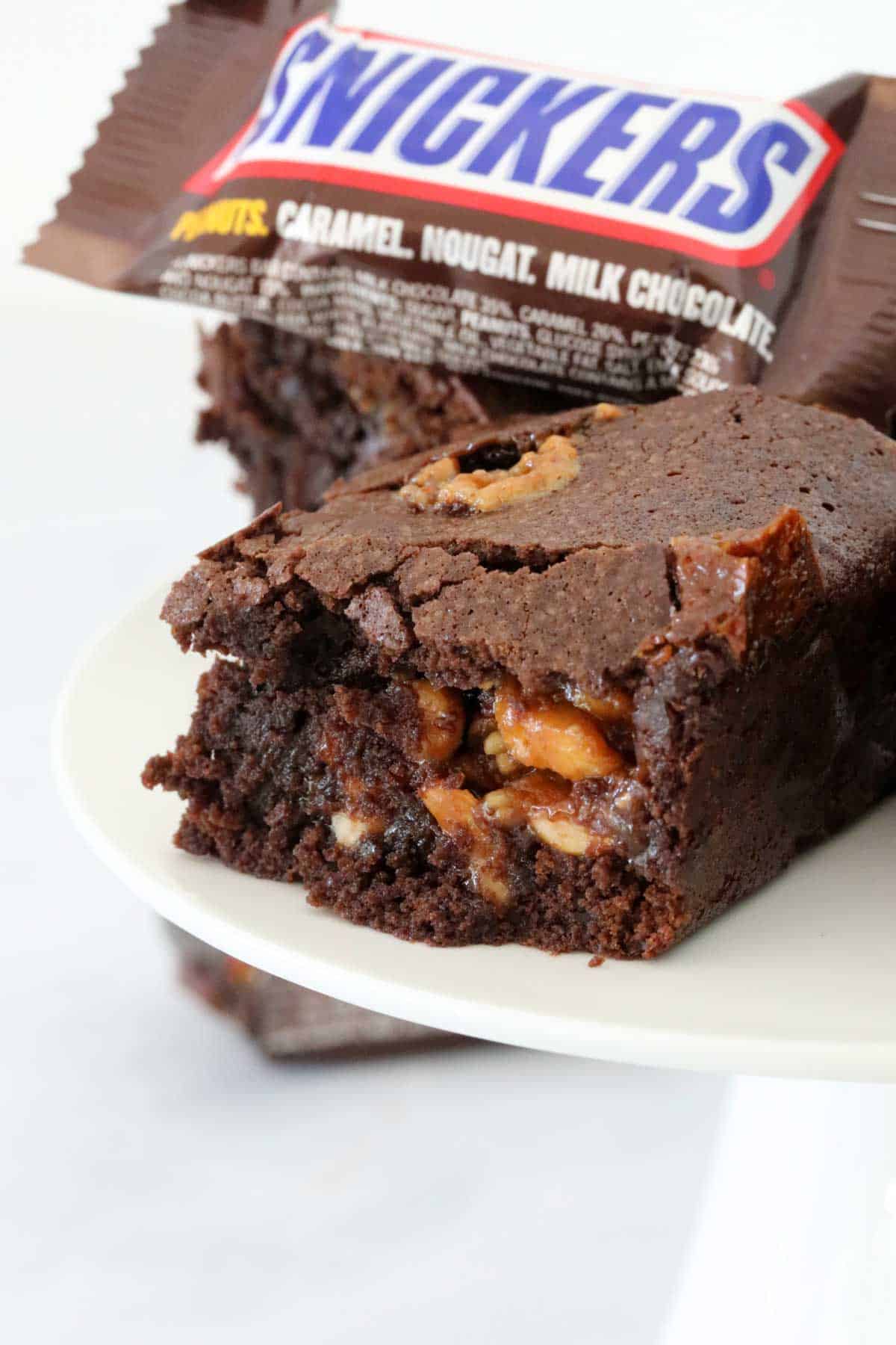 Side view of a square of brownie made with Snickers, with a Snickers chocolate bar in the background.