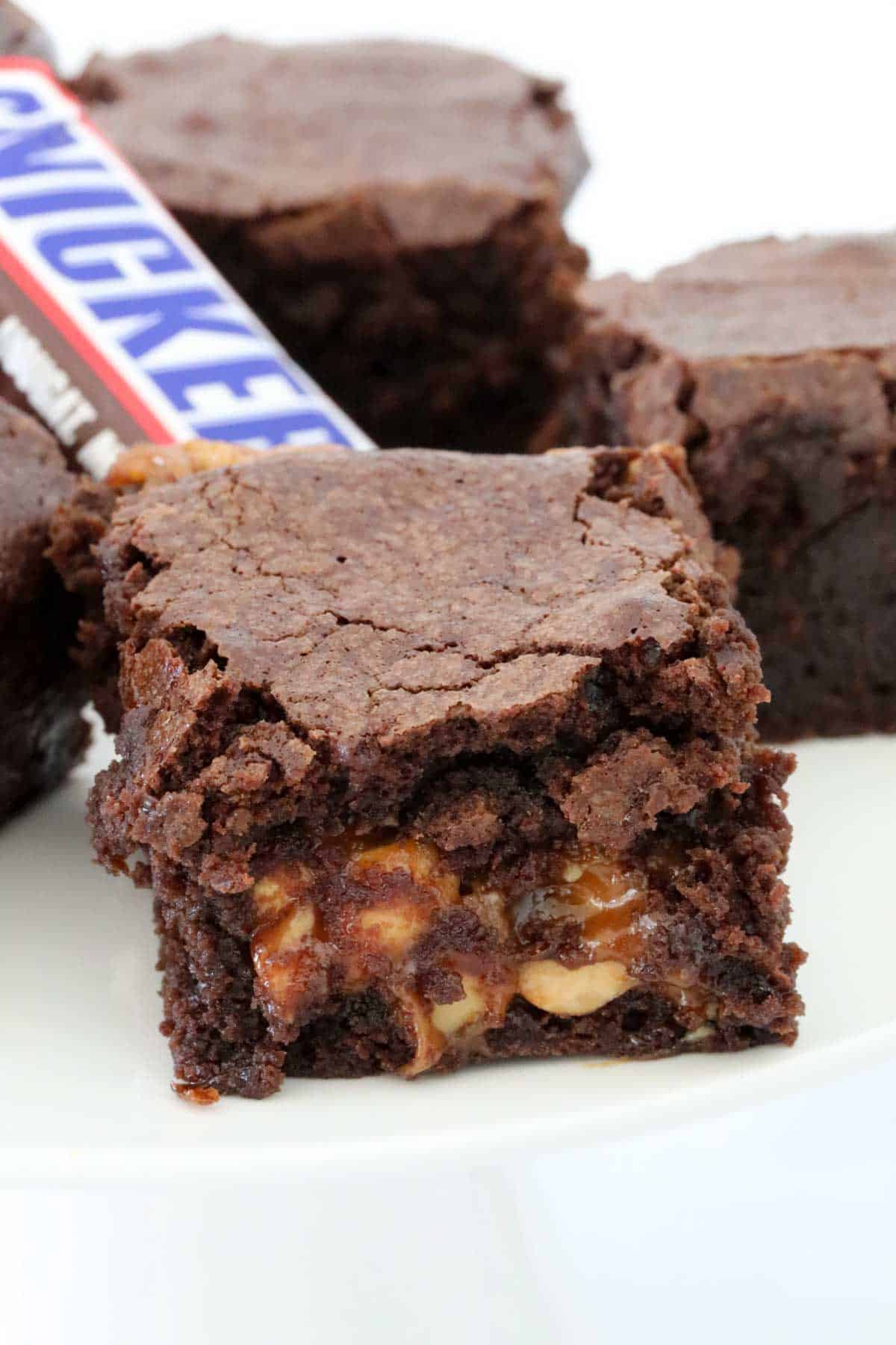 Cut Snickers brownies on a white cake stand, with a Snickers bar just visible in the background.