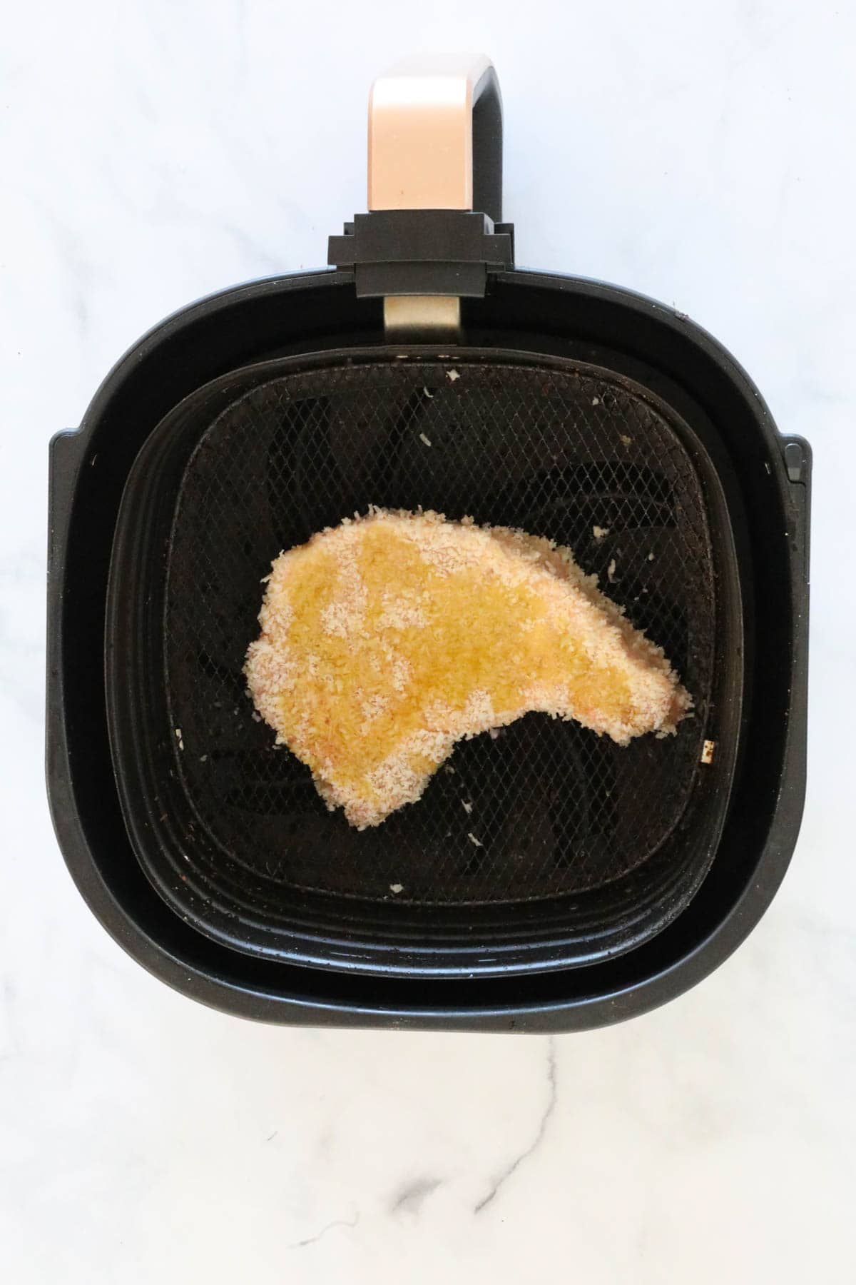 A breaded pork chop in the basket of an air fryer, sprayed with oil.