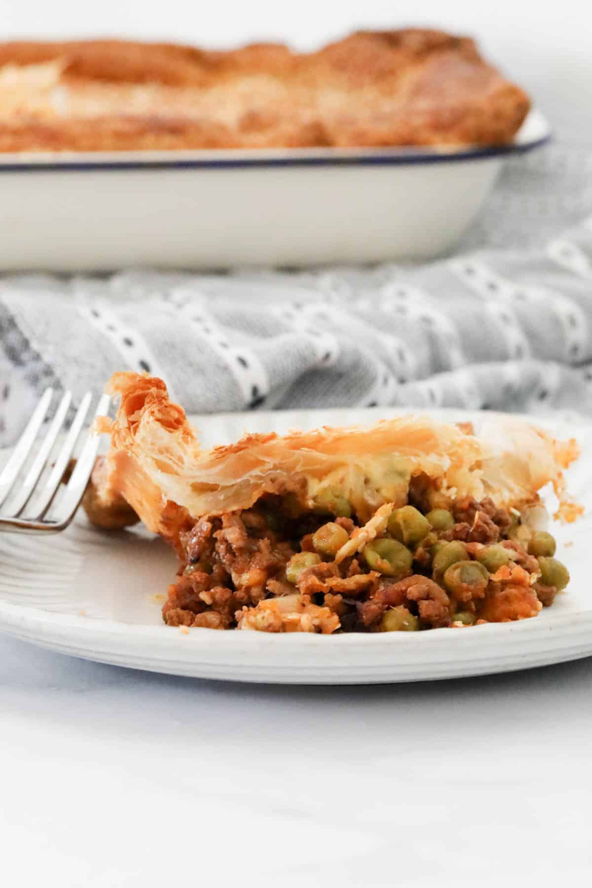 A portion of mince and cheese pie on a white plate, with the baking dish in the background.