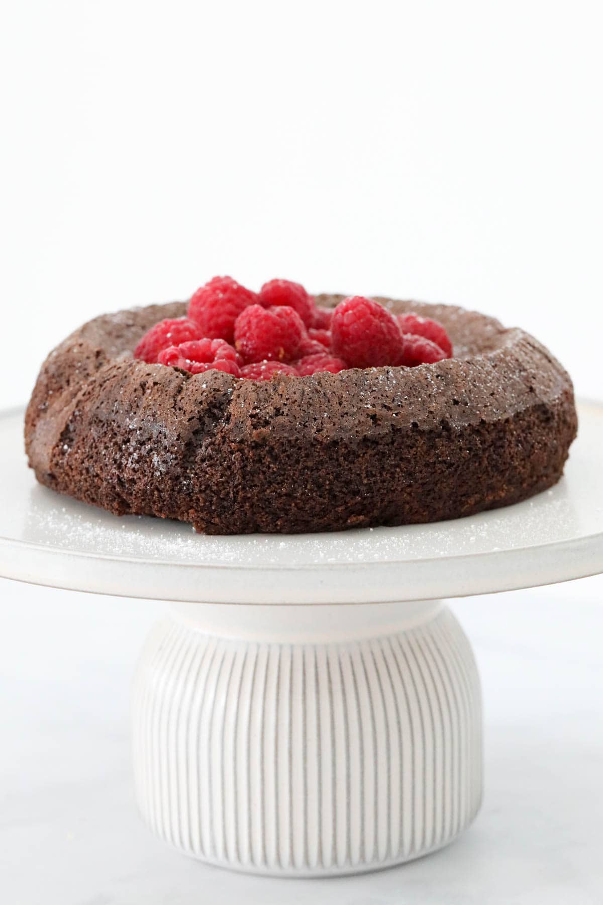 A chocolate olive oil cake topped with fresh raspberries, and served on a white cake stand.