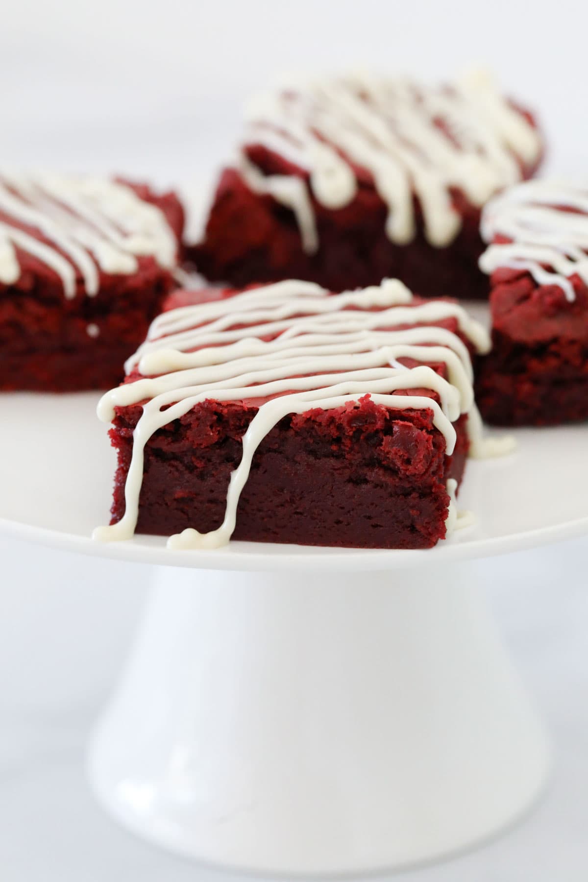 Red velvet brownies drizzled with white chocolate on a white cake stand.