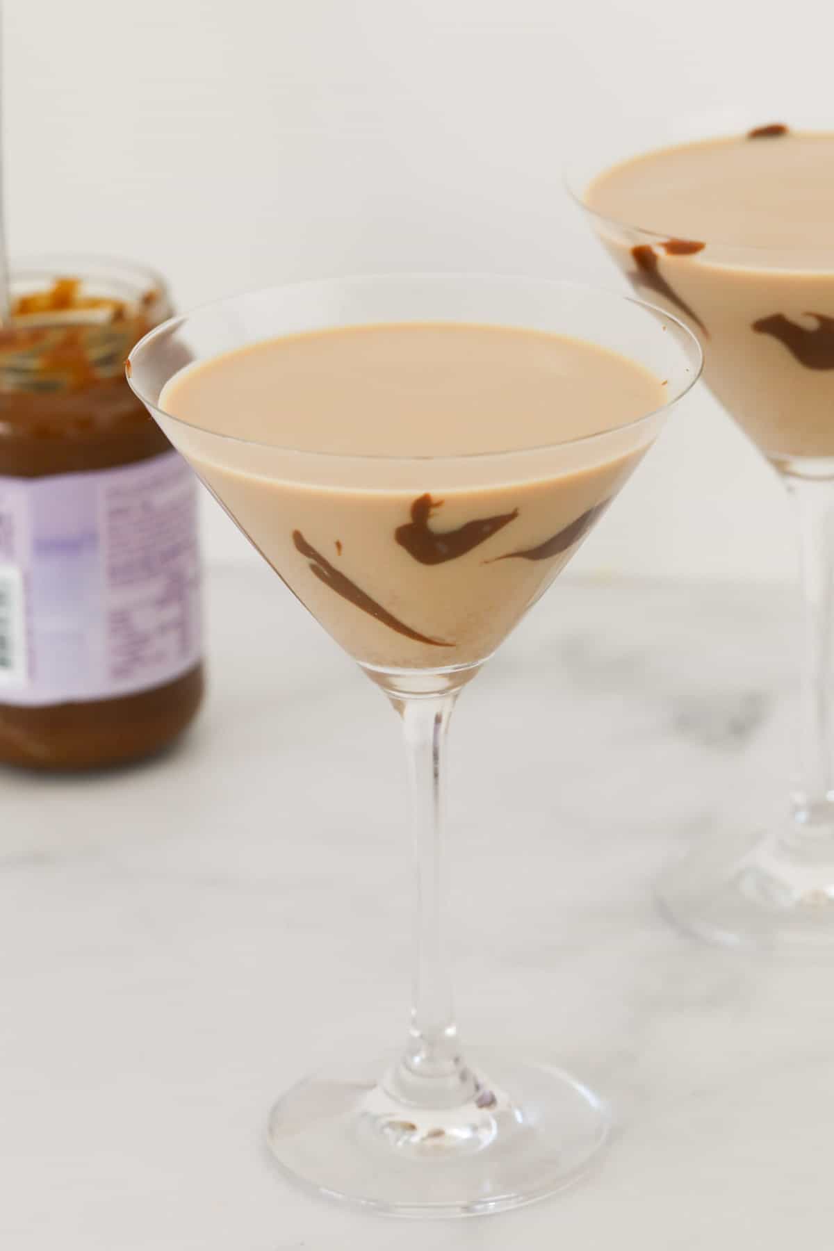 Two glasses of salted caramel cocktail with a jar of caramel sauce just visible behind.