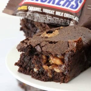 Gooey caramel nut chocolate Snickers brownie with a Snickers bar in the background.