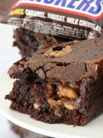 Gooey caramel nut chocolate Snickers brownie with a Snickers bar in the background.