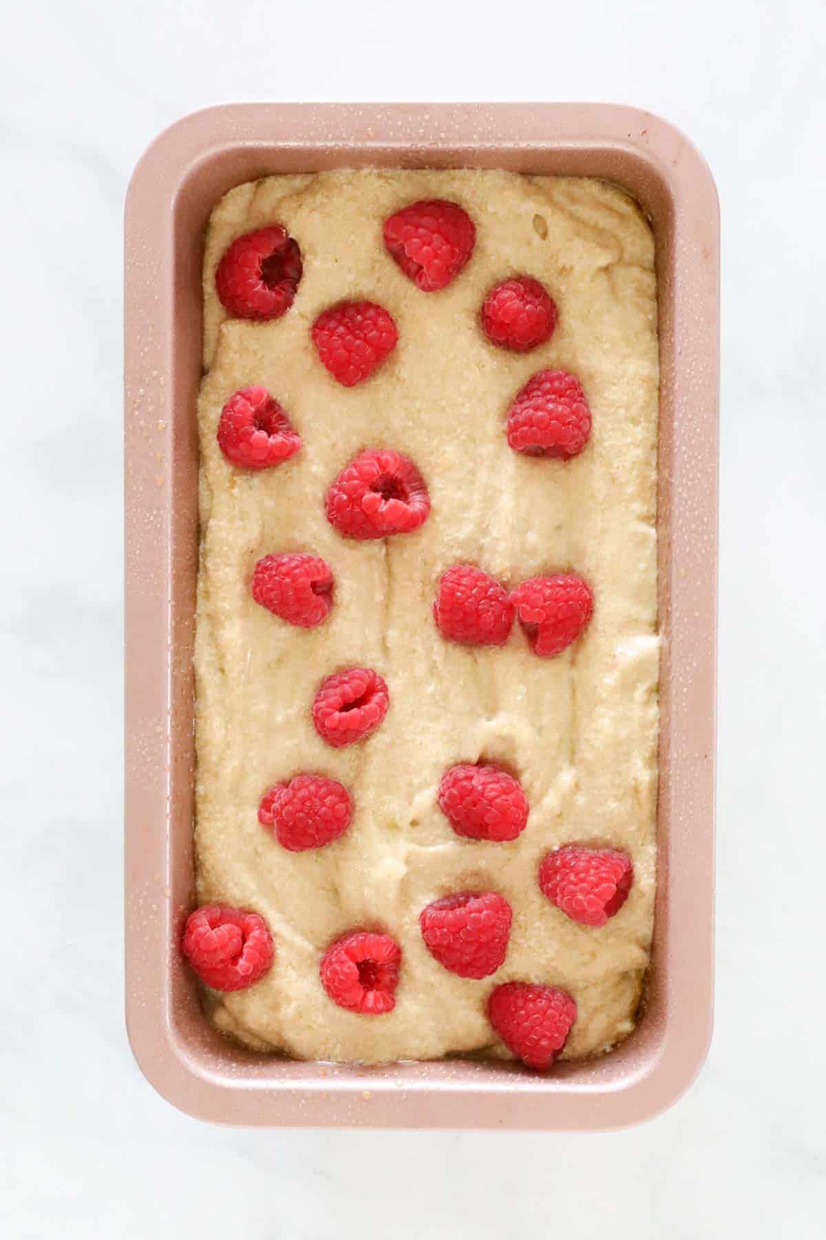 Banana and raspberry loaf mixture in baking tin, with more raspberries placed on top before being baked.