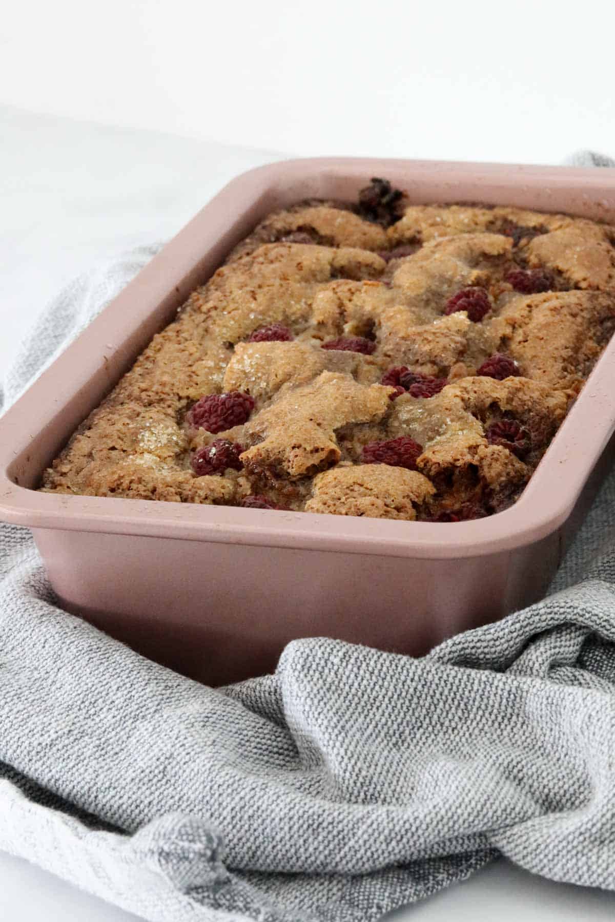 A pale pink baking tin with a baked loaf, dotted with raspberries on top.