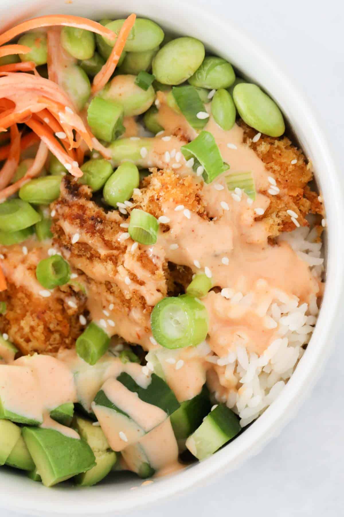 A white bowl of bang bang chicken served with rice and vegetables, and drizzled with bang bang sauce.