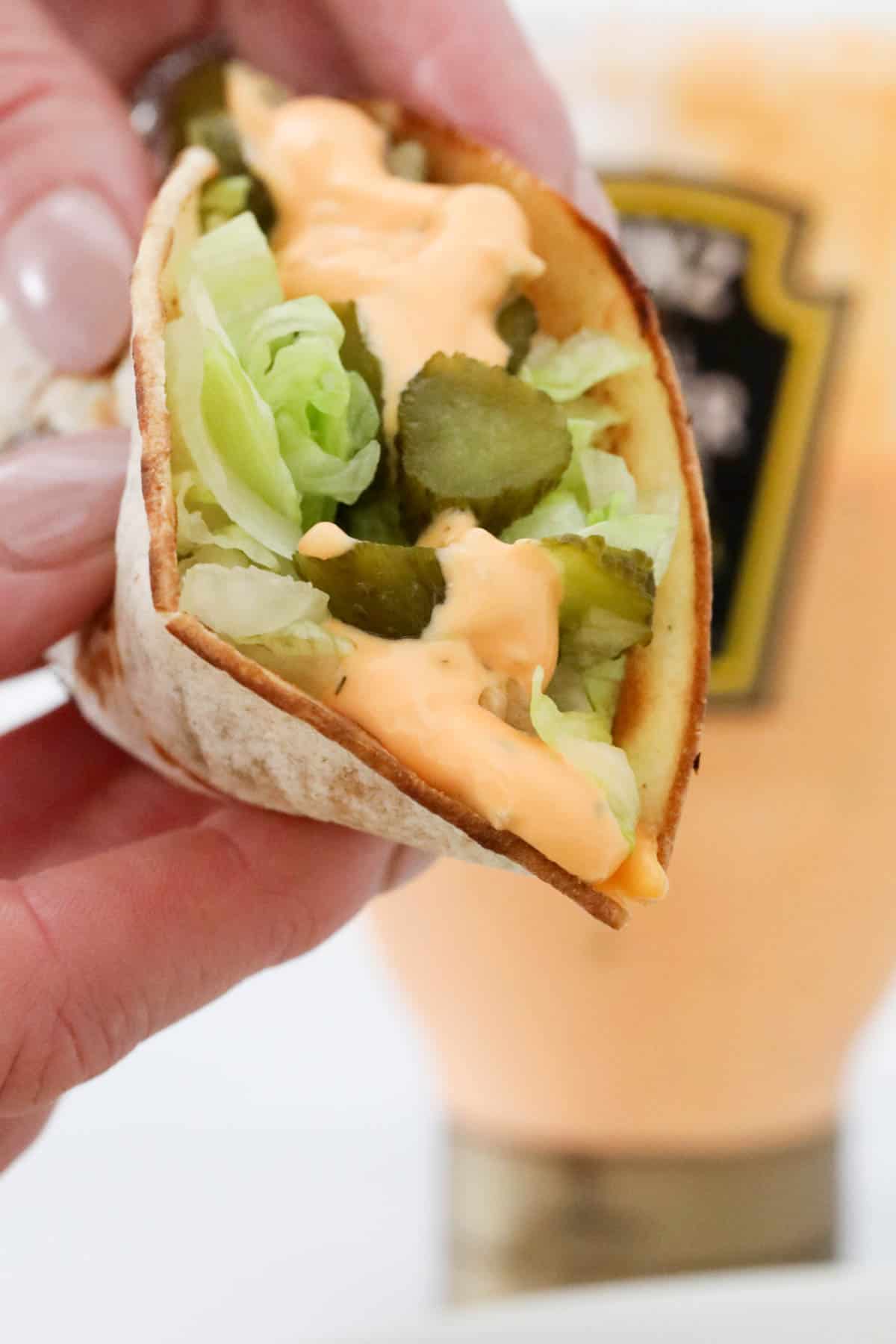 A hand holding a taco with a bottle of burger sauce just visible in the background.