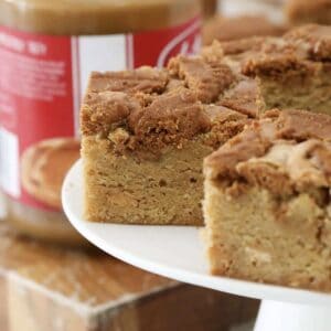 Moist Biscoff Blondies on a white cake stand and with a jar of Biscoff spread in the background.