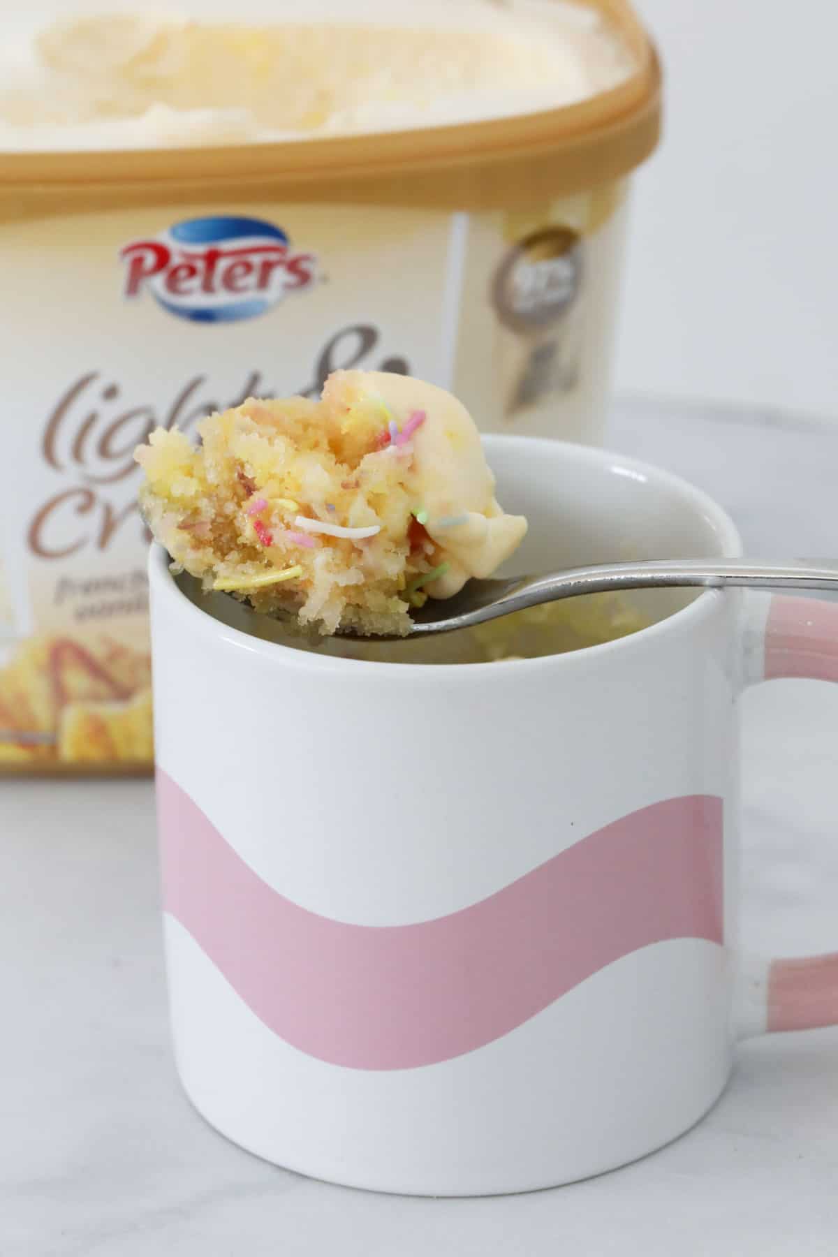 A spoon holding up some vanilla cake with sprinkles, in front of a tub of ice cream.