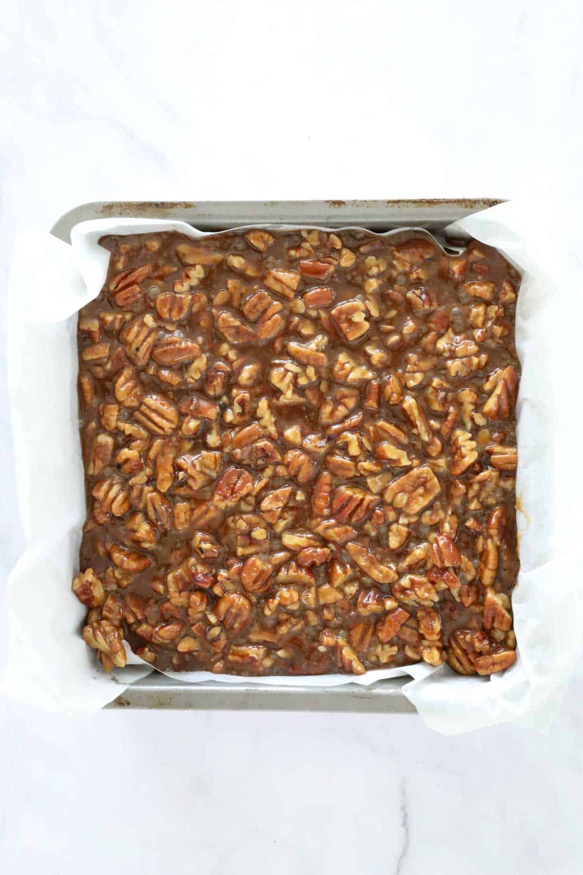 The pecan pie layer poured over the cheesecake layer in the lined tin.