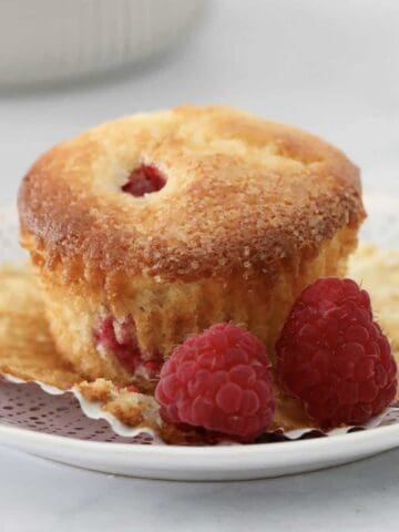 A sweet lemon muffin with raspberries throughout on a plate surrounded by fresh raspberries.