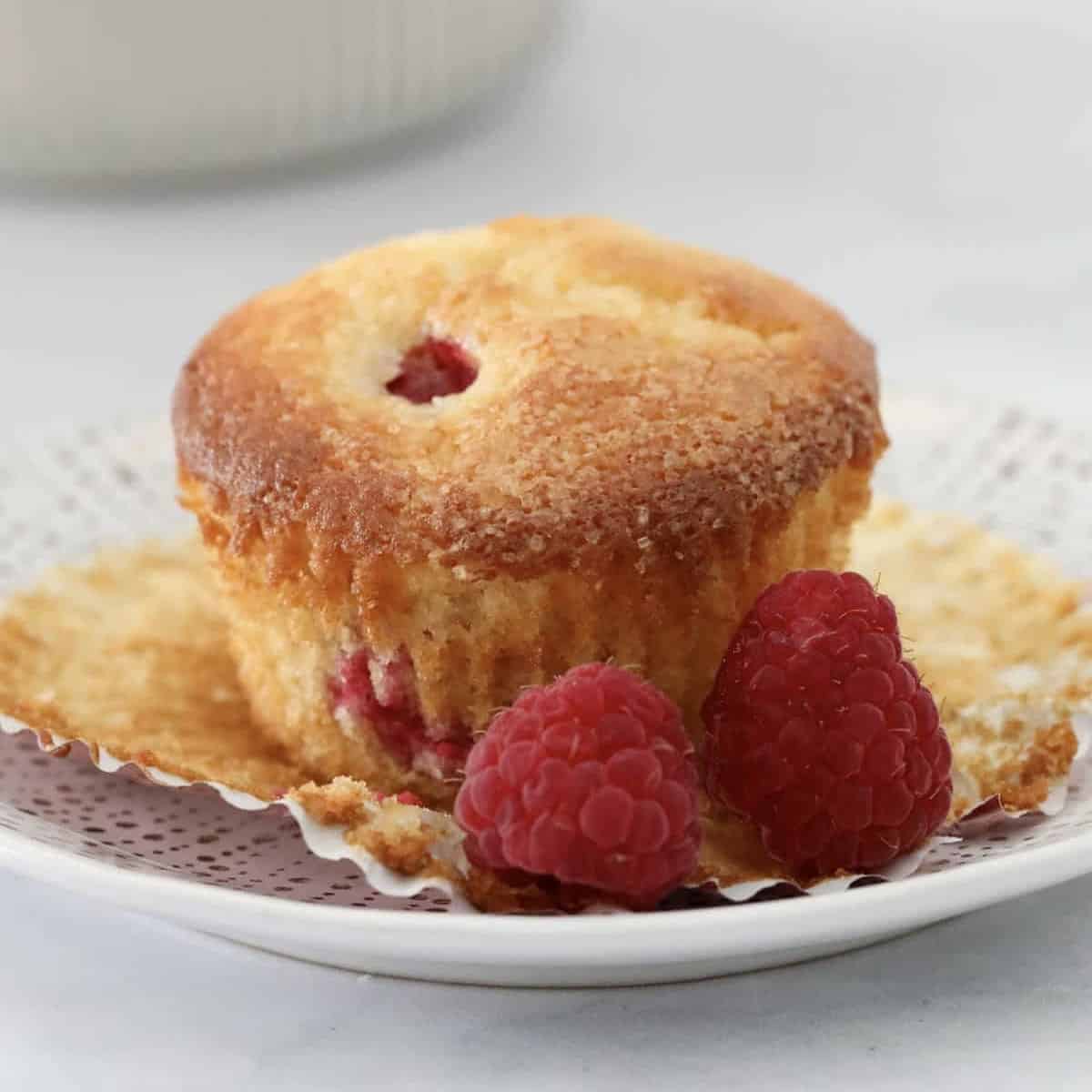 A sweet lemon muffin with raspberries throughout on a plate surrounded by fresh raspberries.