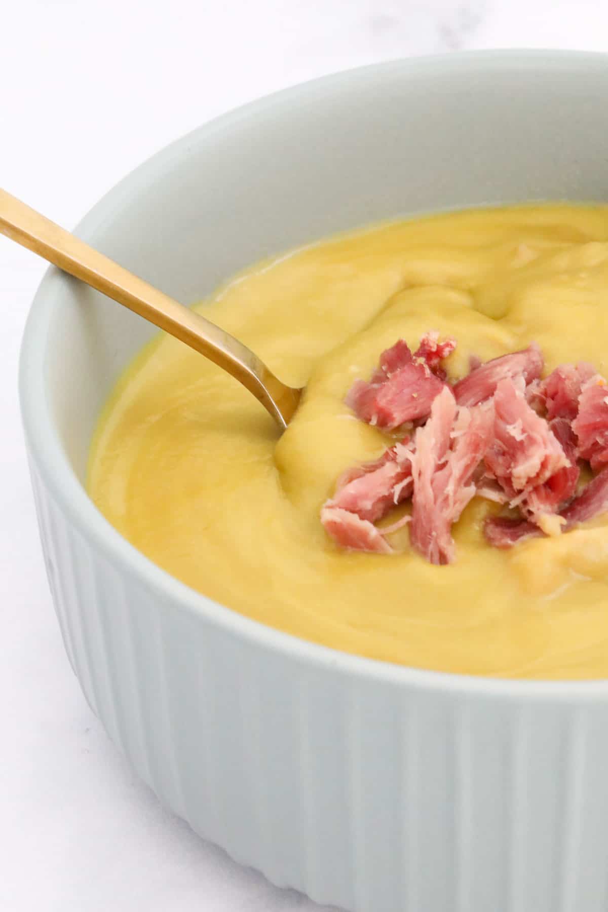 A gold spoon in a bowl of pea soup with pieces of ham hock on top.