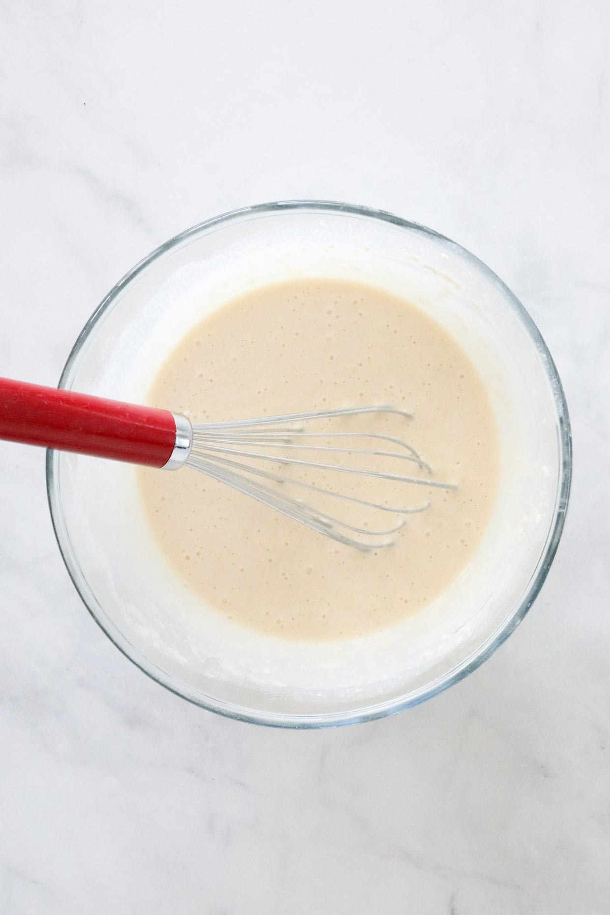 Smooth blinis batter in a glass bowl with a red handled whisk.