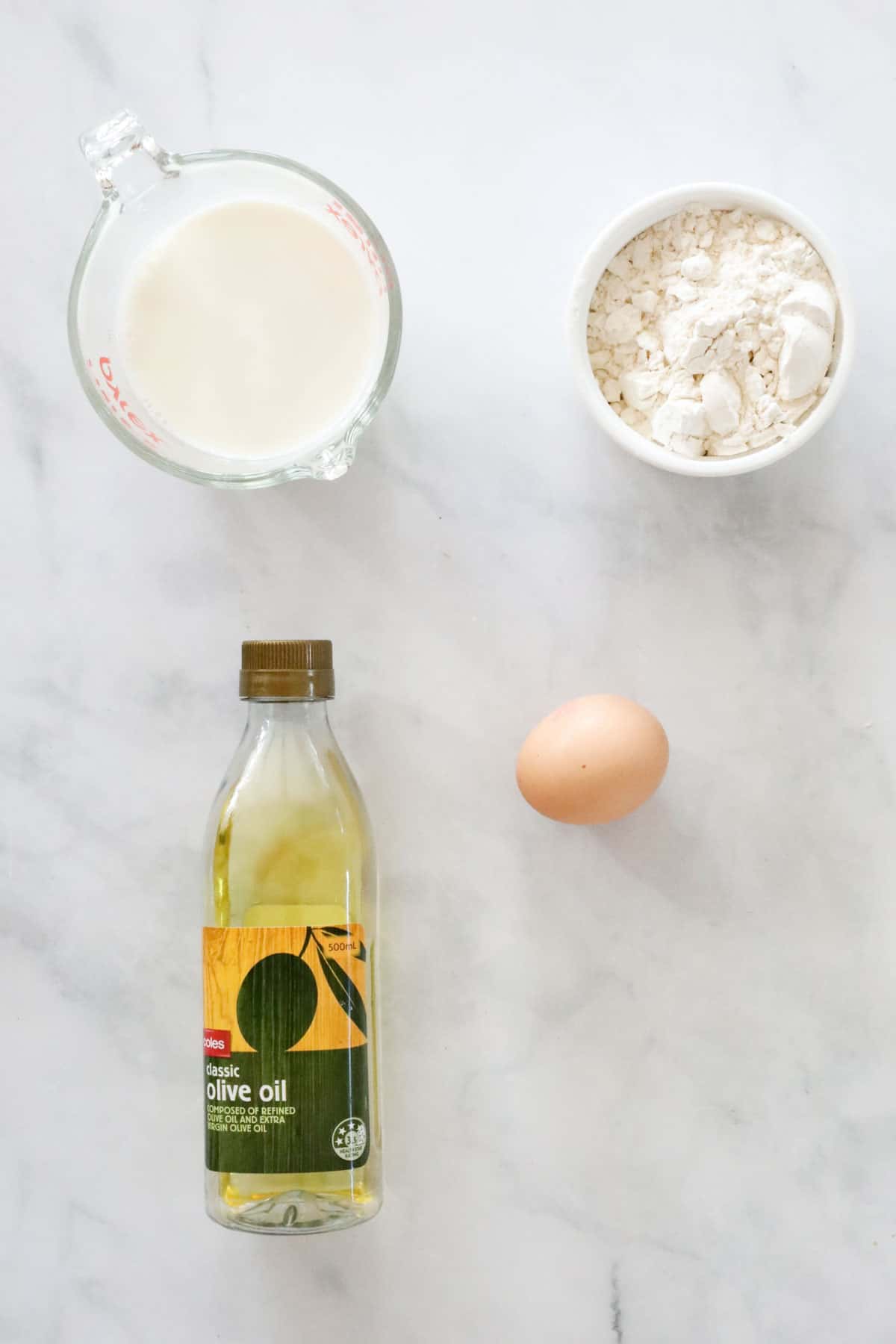 Self-raising flour, milk, an egg and a bottle of olive oil on a bench.