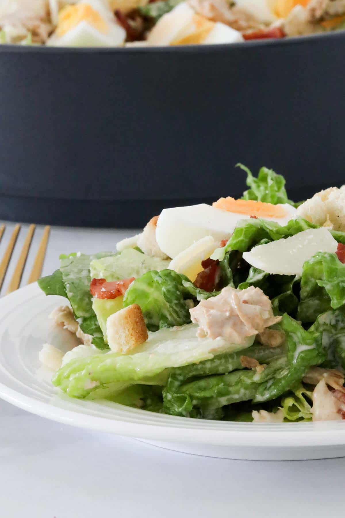 A serving of chicken Caesar salad on a white plate in front of the serving bowl full of salad.