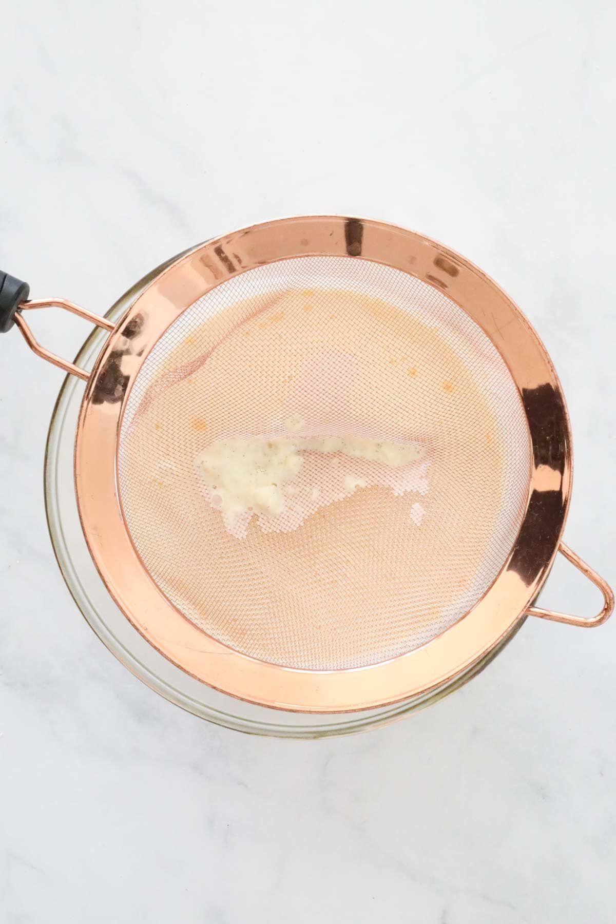 The cream mixture being poured through a sieve onto the egg yolk mixture.