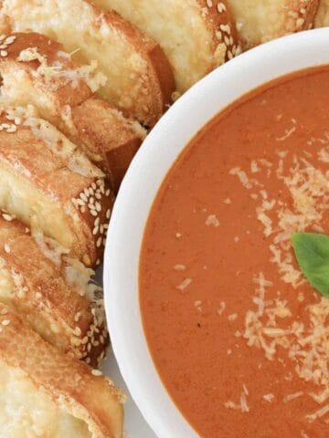 A bowl of roasted tomato soup with grated parmesan, basil and cheesy bread on the side.