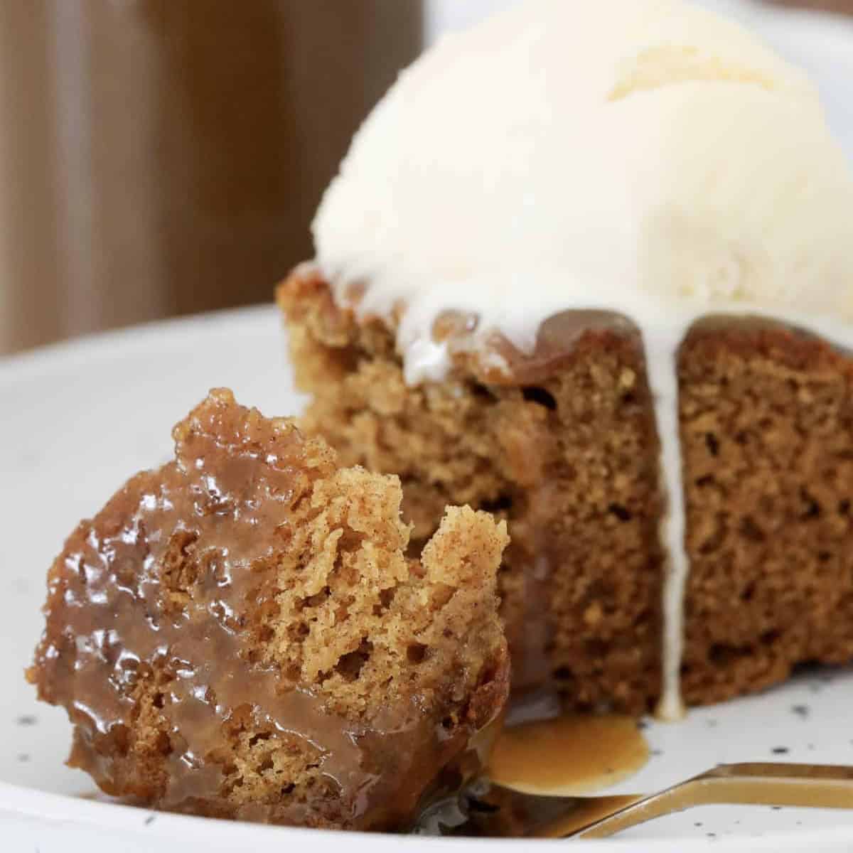 A forkful of moist sticky date pudding with caramel sauce and a scoop of vanilla ice cream.