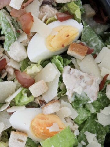 A bowl filled with chicken caesar salad with bacon, parmesan, egg and croutons.