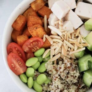 A bowl filled with quinoa, chicken, edamame, almonds, sweet potato, cucumber and tomatoes.