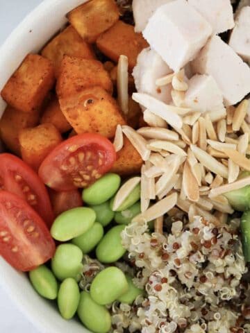 A bowl filled with quinoa, chicken, edamame, almonds, sweet potato, cucumber and tomatoes.