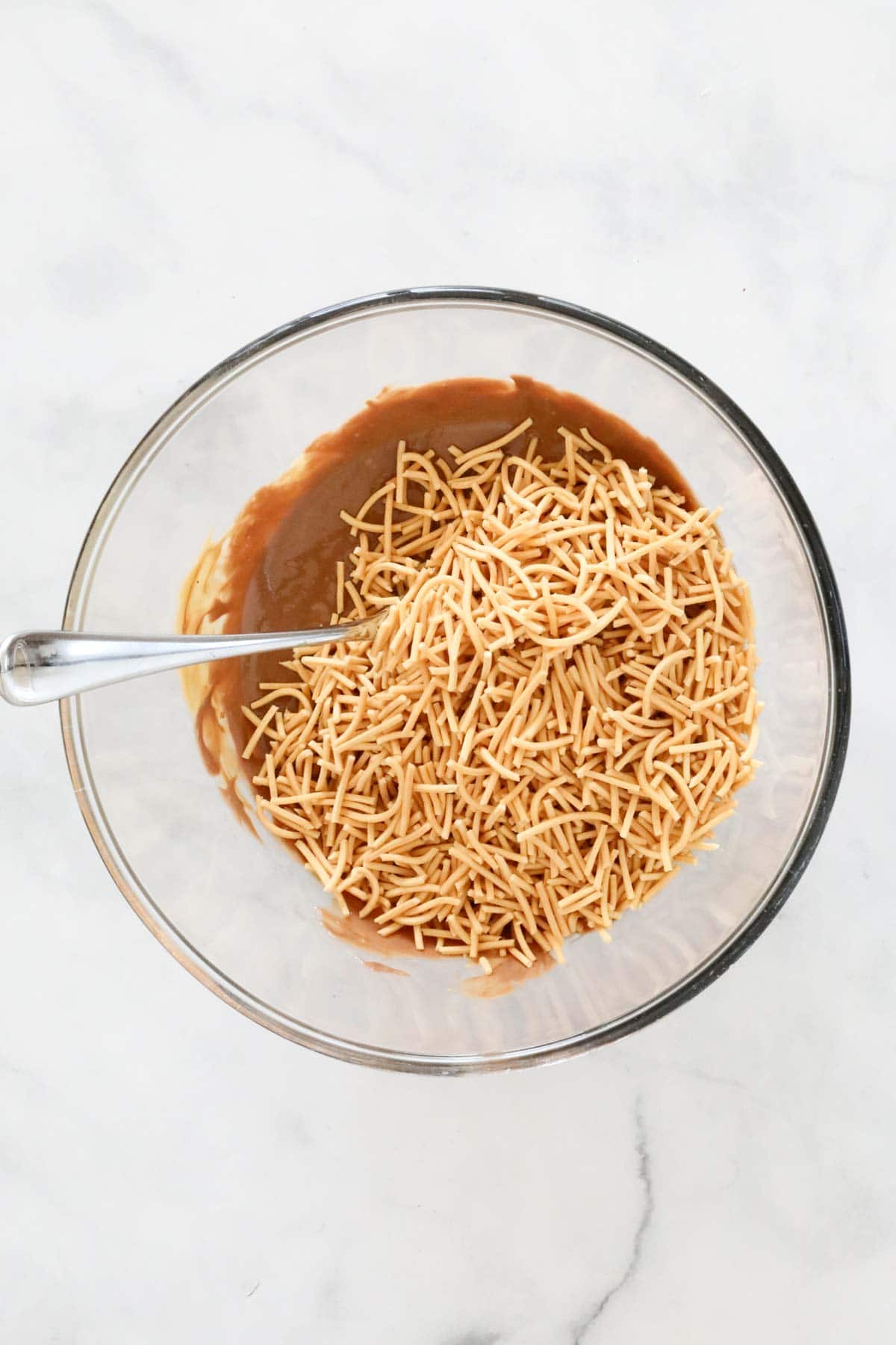 Fried noodles added to the melted mixture in a glass bowl.