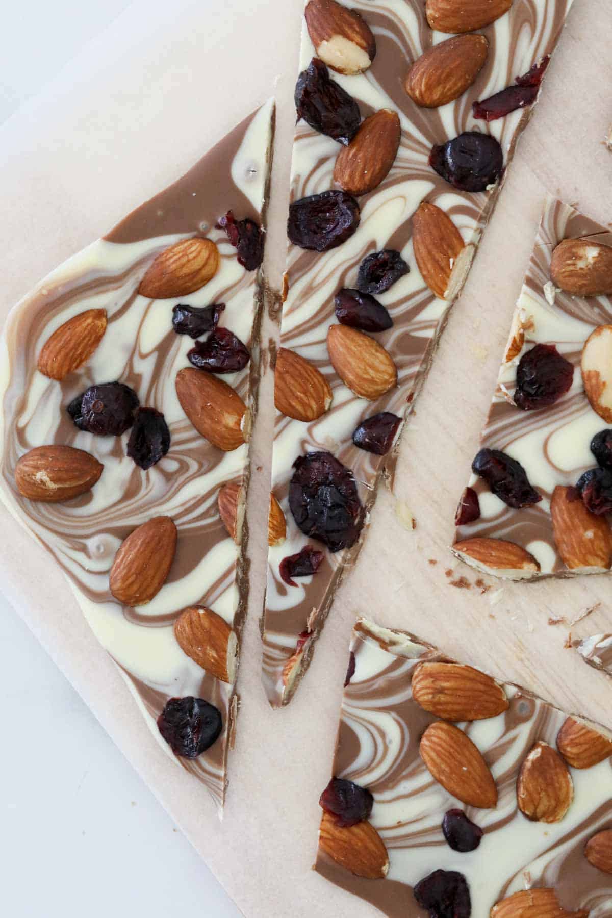 A slab of milk and white chocolate bark with cranberries and almonds, cut into pieces on a bench.