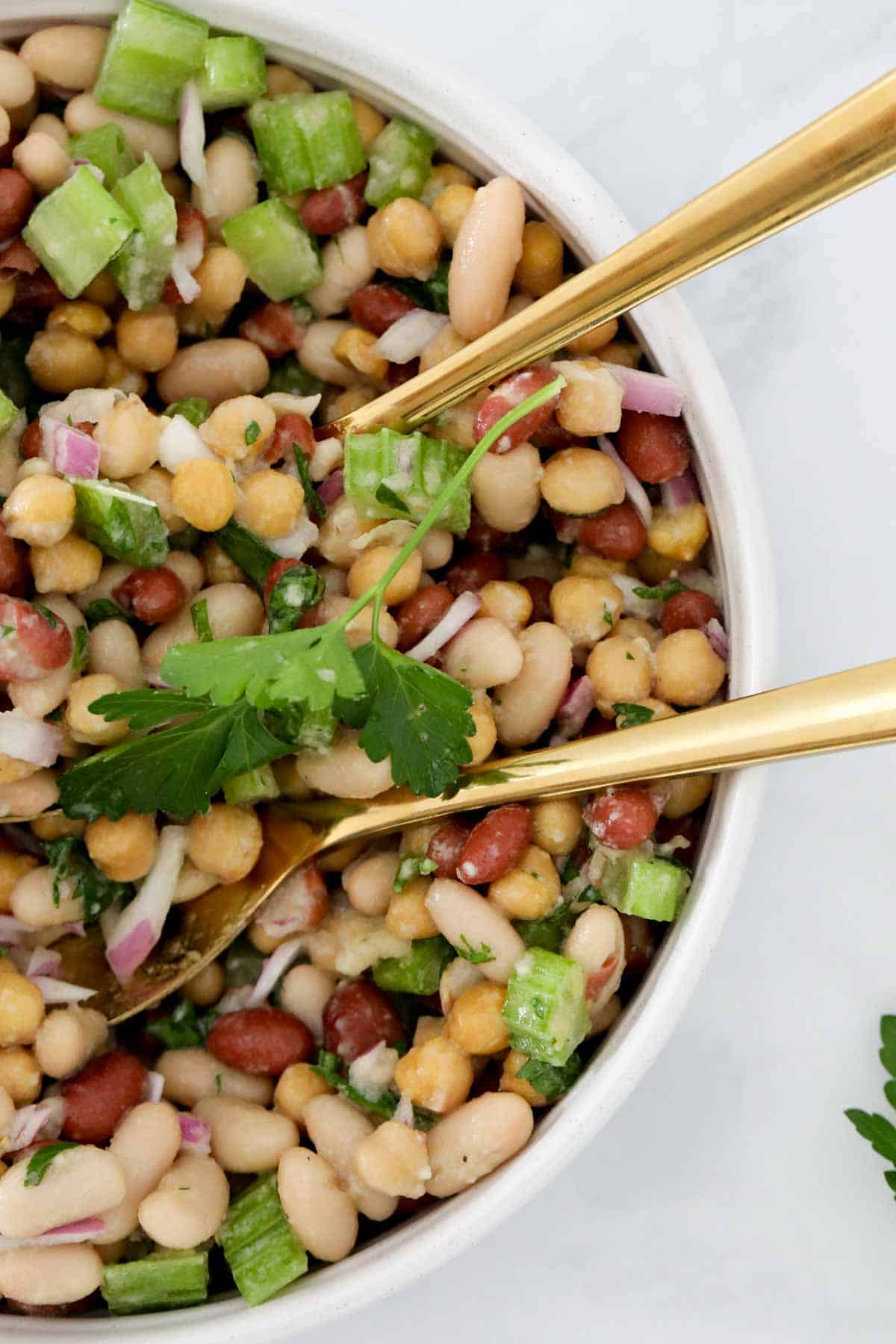 Gold salad servers placed in a bowl of bean and chickpea salad.